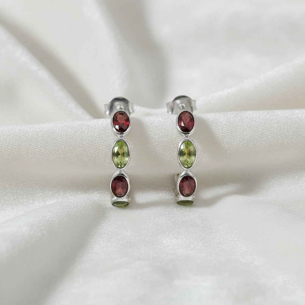 Silver hoop earrings featuring oval red garnet and green peridot stones on an off white velvet fabric close up shot
