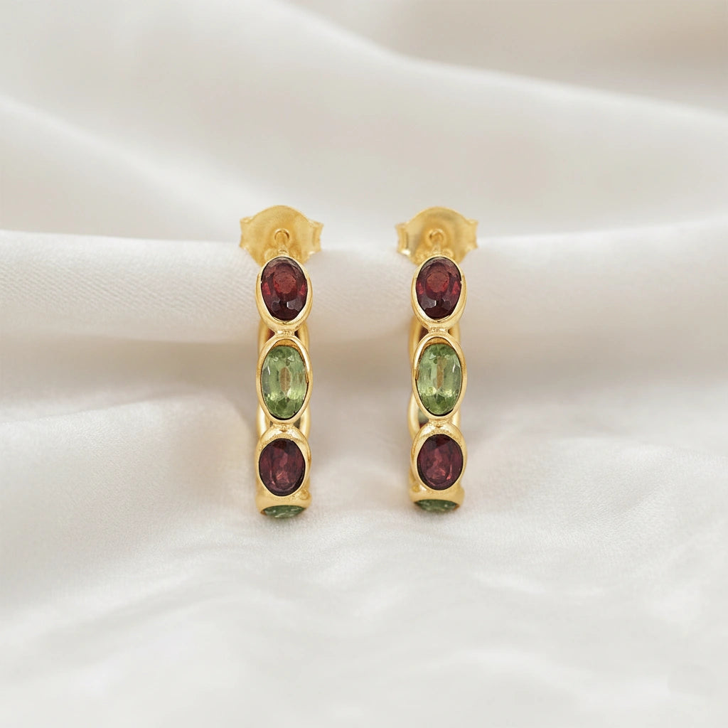 Gold plated hoop earrings featuring oval cut red garnet and green peridot stones on an off white velvet fabric close up angle shot