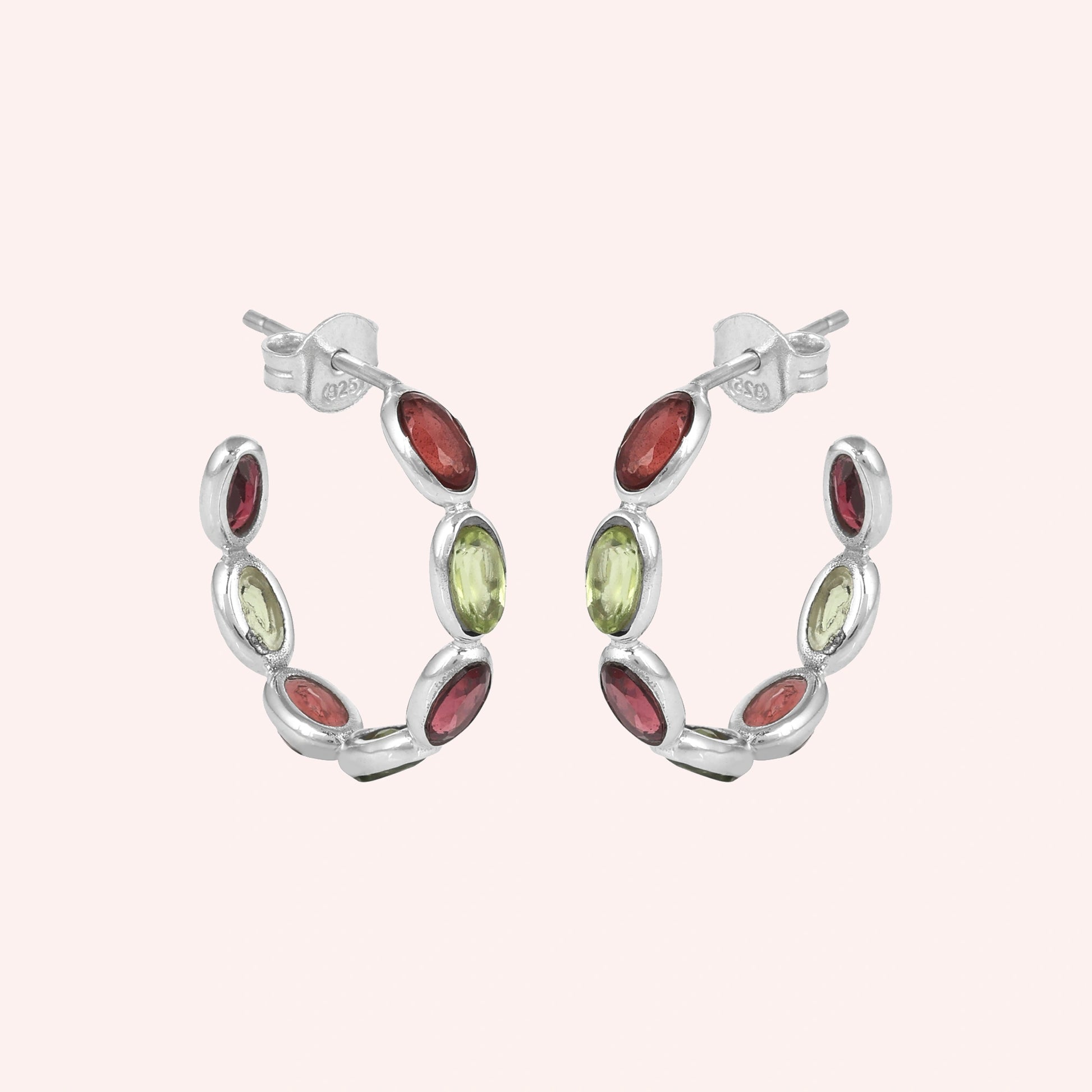 Silver hoop earrings featuring oval cut red garnet and green peridot stones on a pink background studio shot angled view
