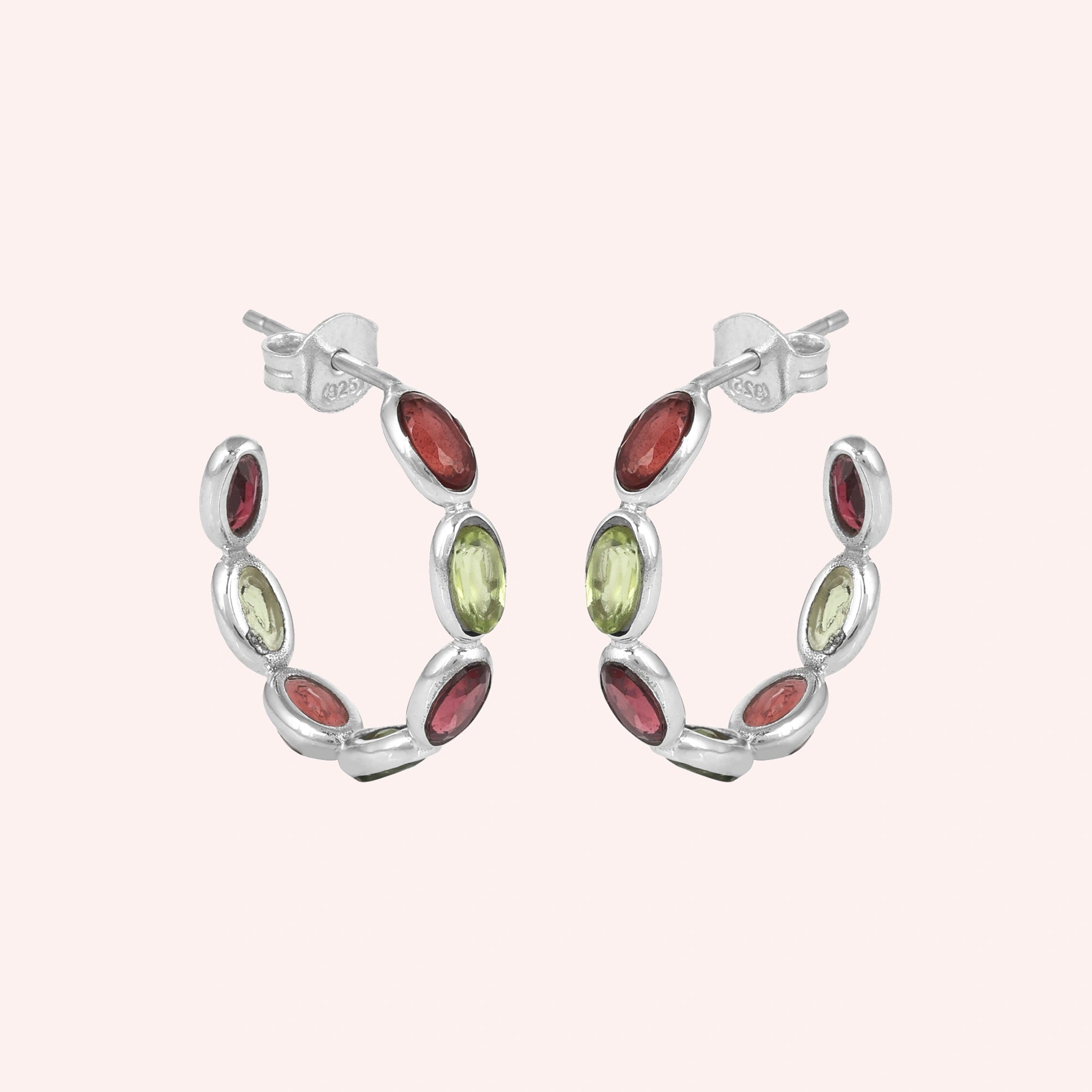 Silver hoop earrings featuring oval cut red garnet and green peridot stones on a pink background studio shot angled view