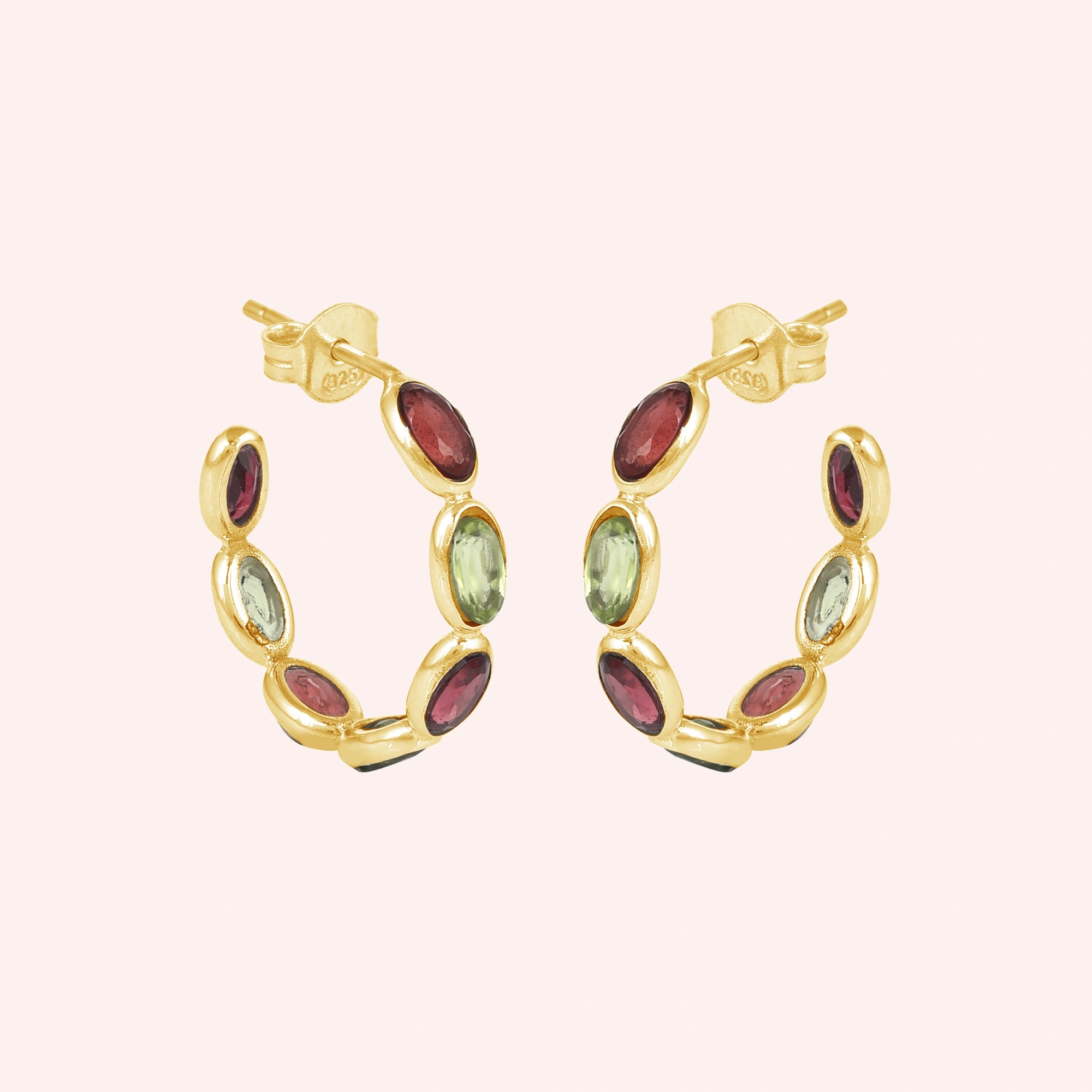 Gold plated hoop earrings featuring oval cut red garnet and green peridot stones on a pink background angled view shot