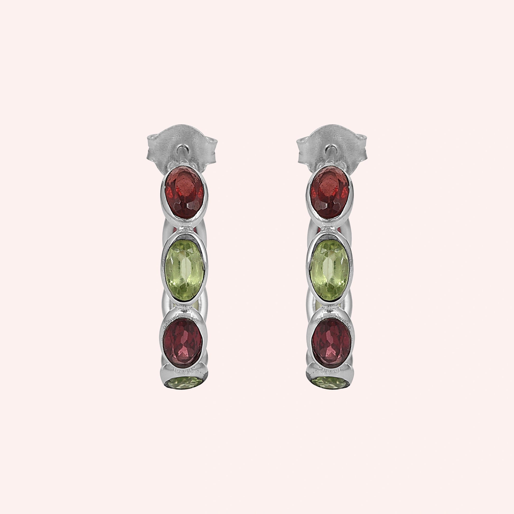 Silver hoop earrings featuring oval cut red garnet and green peridot stones on a pink background studio shot top down angle