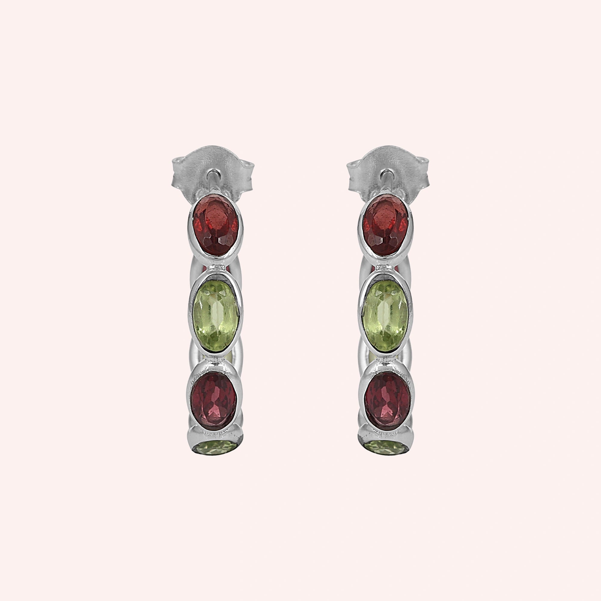 Silver hoop earrings featuring oval cut red garnet and green peridot stones on a pink background studio shot top down angle