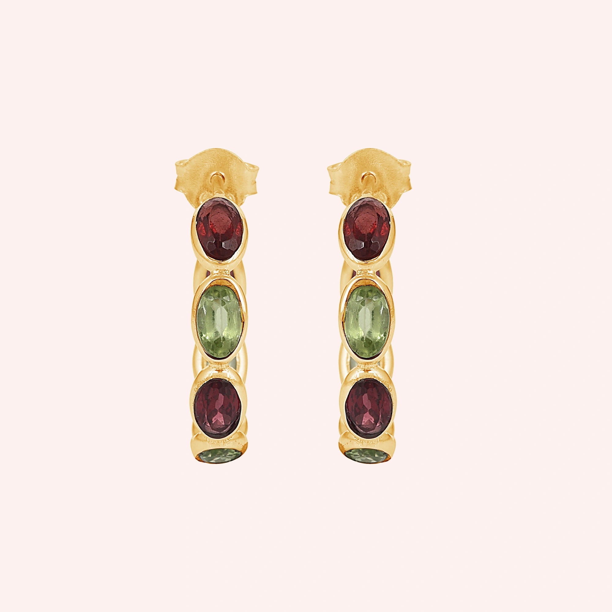 Gold plated hoop earrings featuring oval cut red garnet and green peridot stones on a pink background top down angle shot 