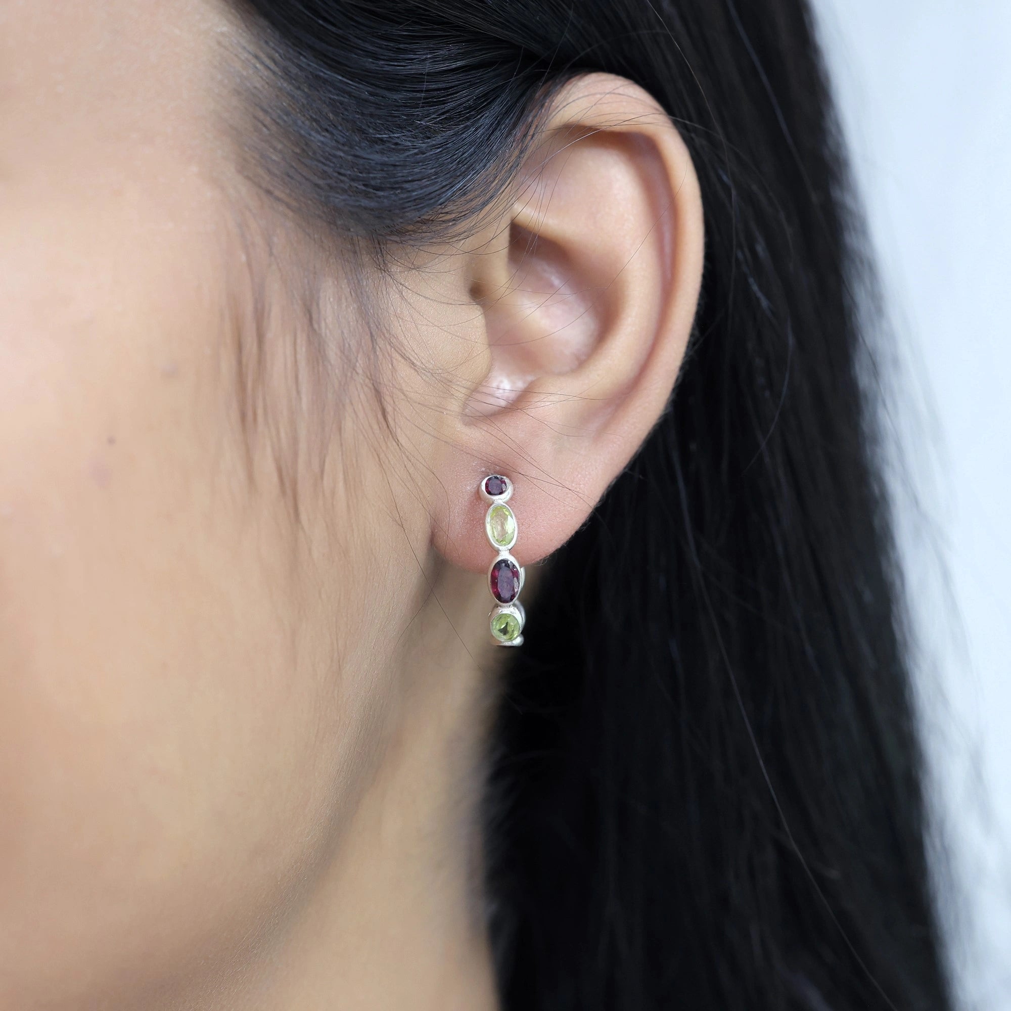 Side angle view of silver peridot hoop earrings featuring red stones worn on the ear with background texture view