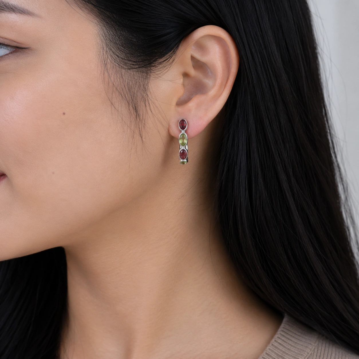 Side angle view of silver peridot hoop earrings featuring red stones worn on the ear with background texture view