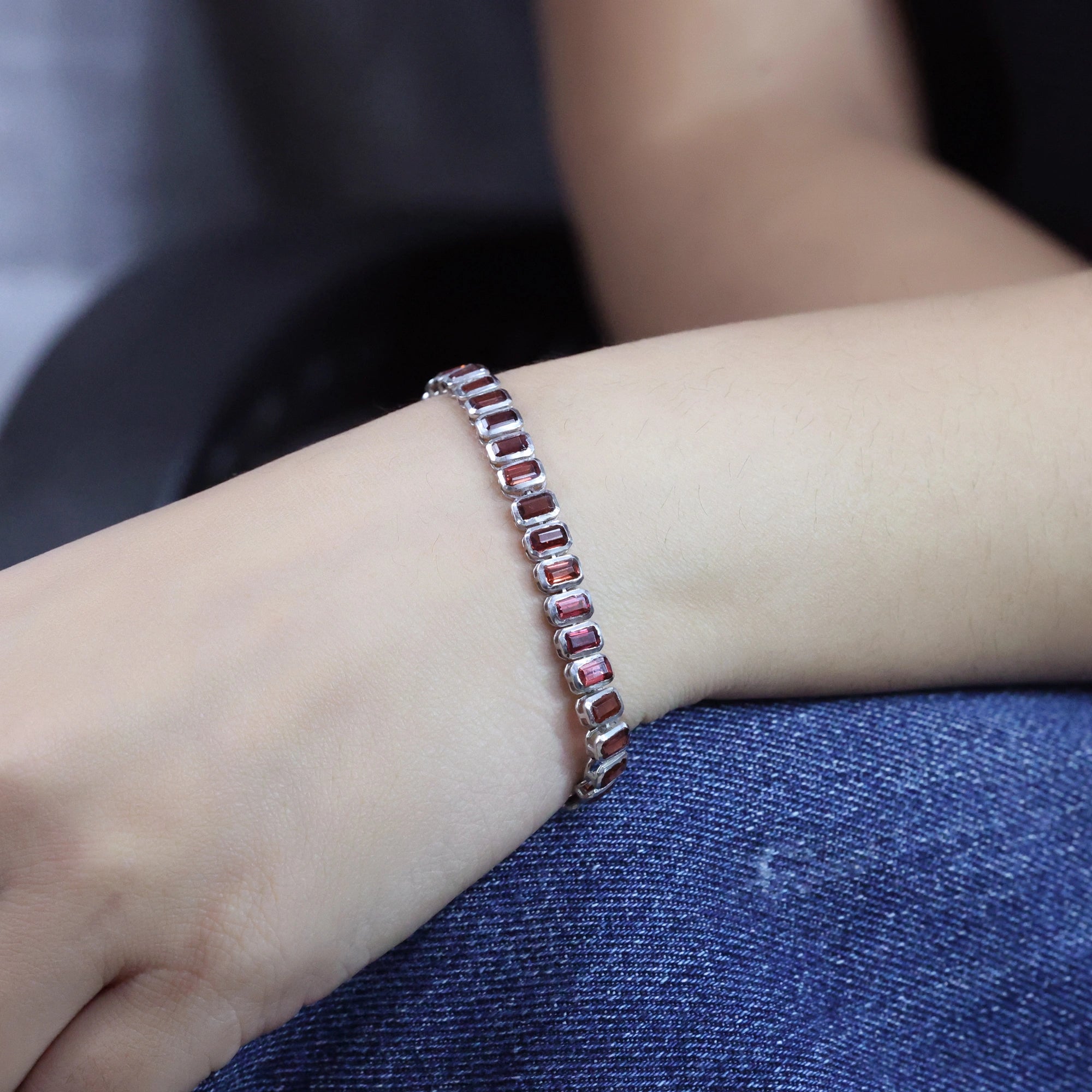 Angled view of tennis bracelet with red garnet baguette stones worn on a wrist over blue denim jeans background texture