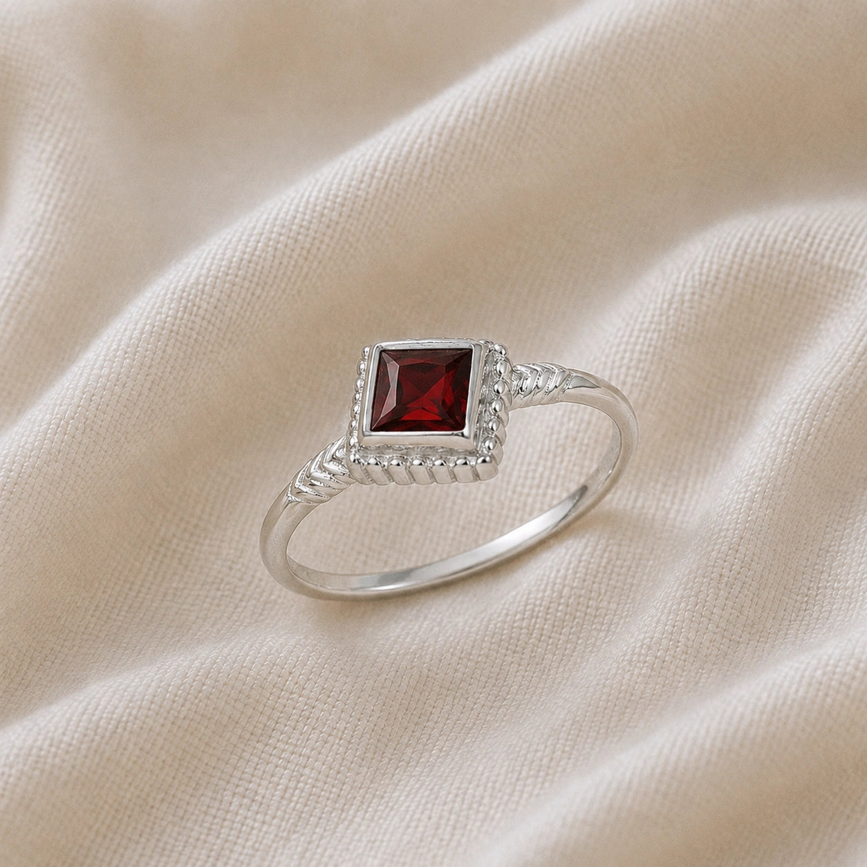 Princess_cut_ring_sterling_silver_with_square_garnet_and_detailed_band_slightly_tilted_view_on_soft_fabric_background