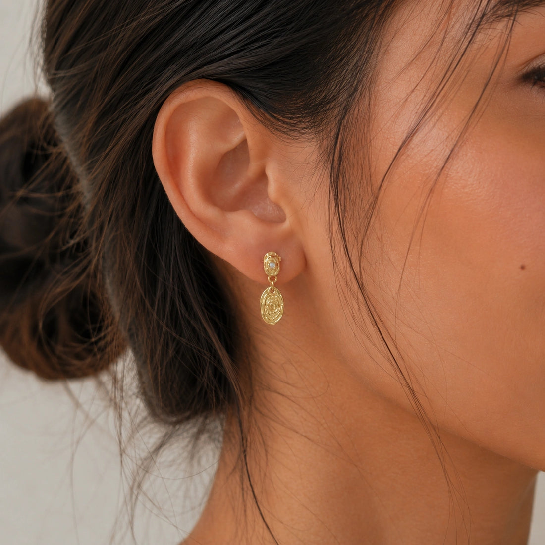 Gold_dangle_earrings_with_gold_plated_silver_textured_oval_charms_and_moonstone_at_front_angle_on_soft_fabric_background