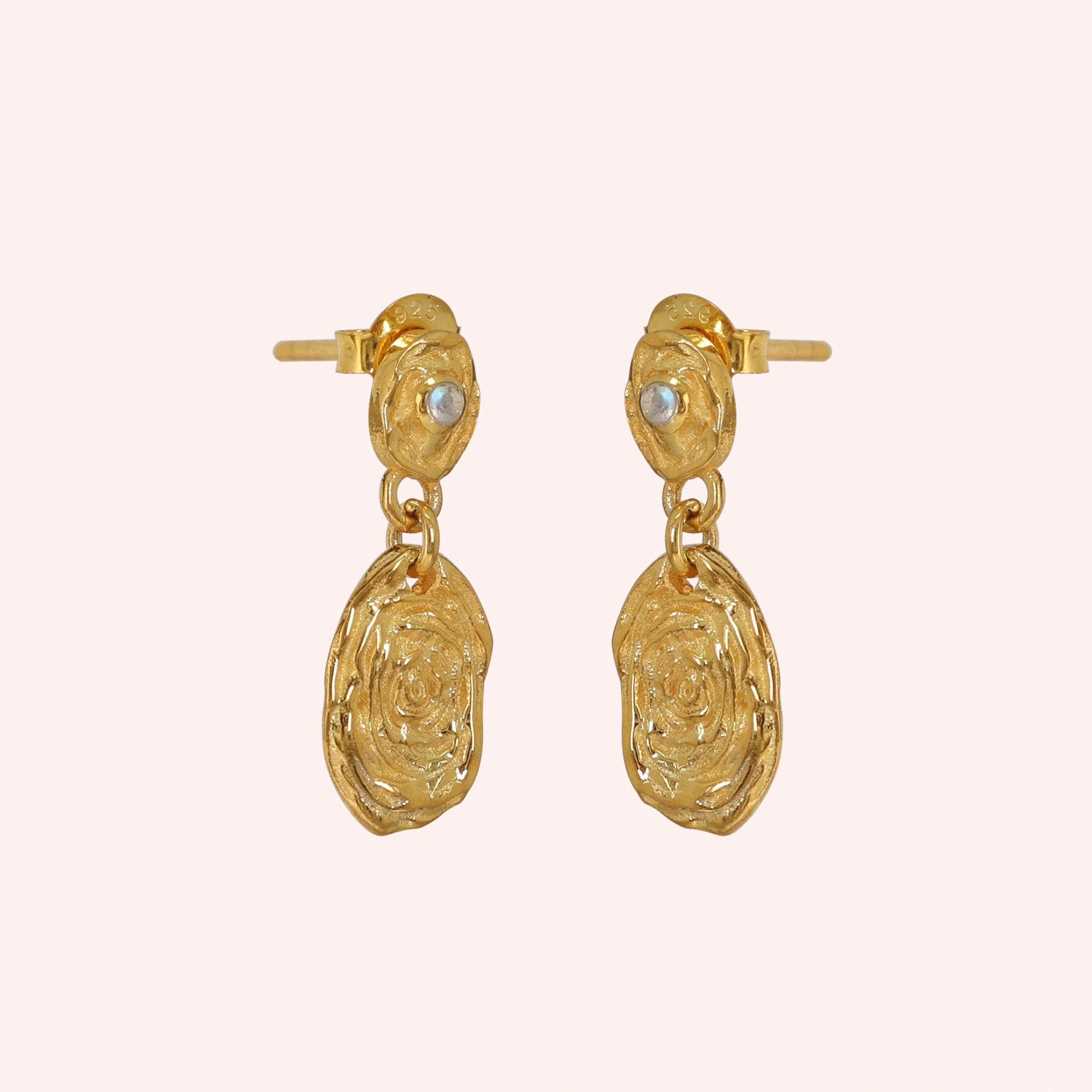 Gold_dangle_earrings_with_gold_plated_silver_textured_oval_charms_and_moonstone_at_side_angle_on_solid_pink_background