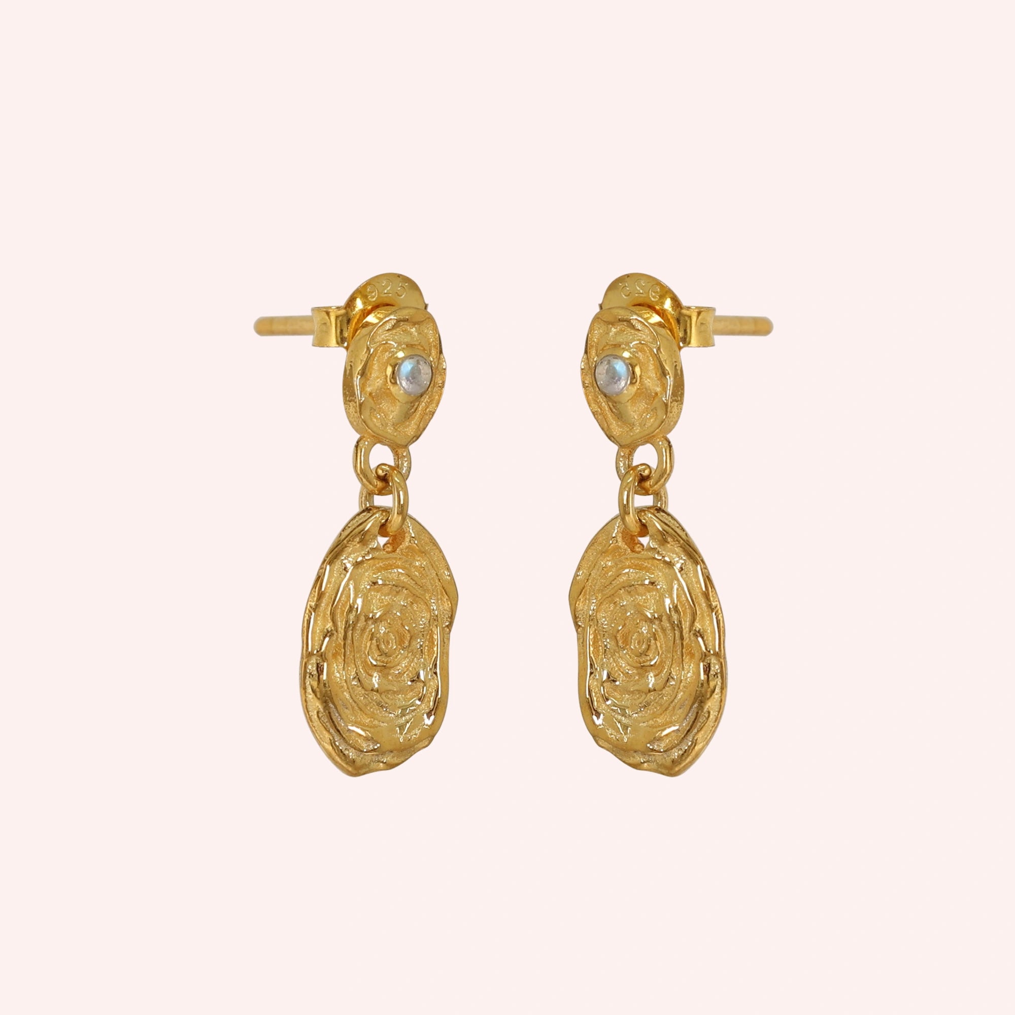 Gold_dangle_earrings_with_gold_plated_silver_textured_oval_charms_and_moonstone_at_side_angle_on_solid_pink_background