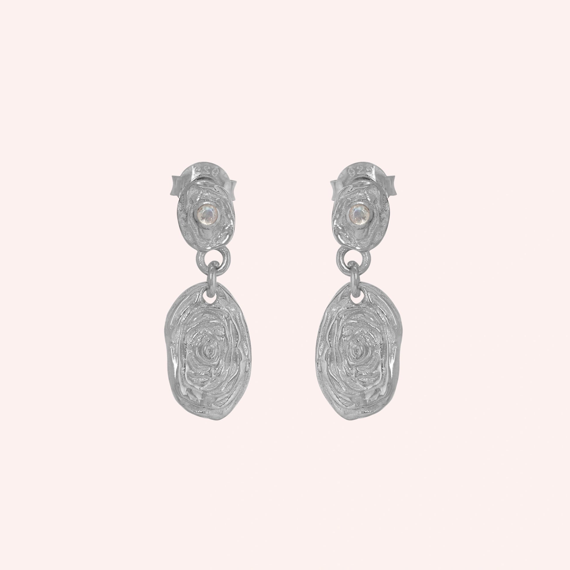 Gold_dangle_earrings_with_sterling_silver_textured_oval_charms_and_moonstone_at_front_angle_on_solid_pink_background