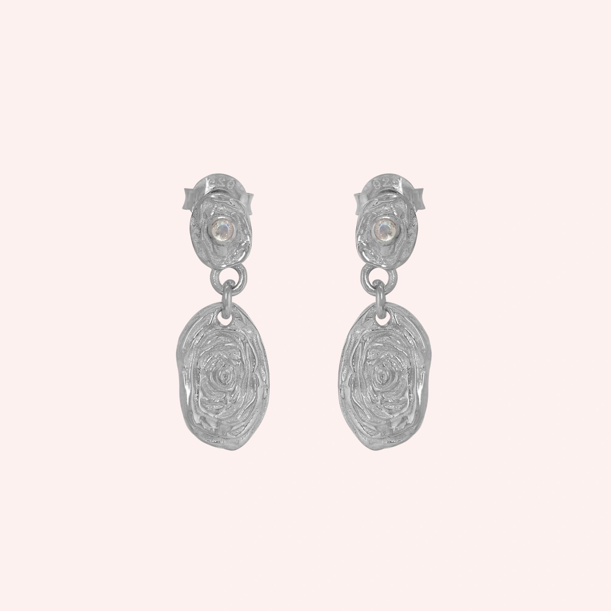 Gold_dangle_earrings_with_sterling_silver_textured_oval_charms_and_moonstone_at_front_angle_on_solid_pink_background