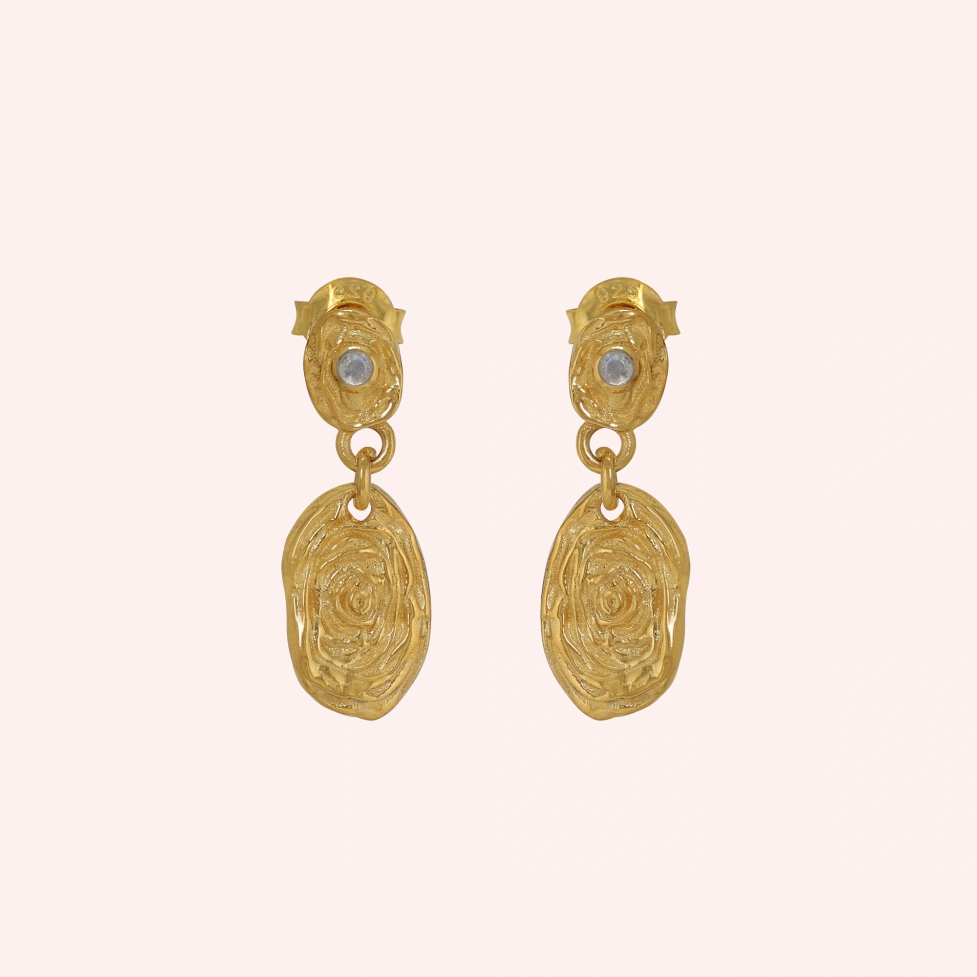 Gold_dangle_earrings_with_gold_plated_silver_textured_oval_charms_and_moonstone_at_front_angle_on_solid_pink_background