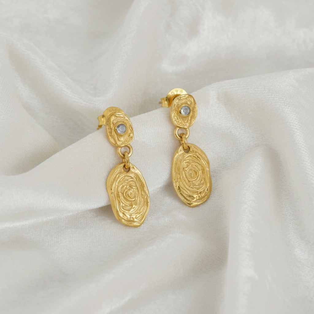 Gold_dangle_earrings_with_gold_plated_silver_textured_oval_charms_and_moonstone_at_front_angle_on_soft_fabric_background
