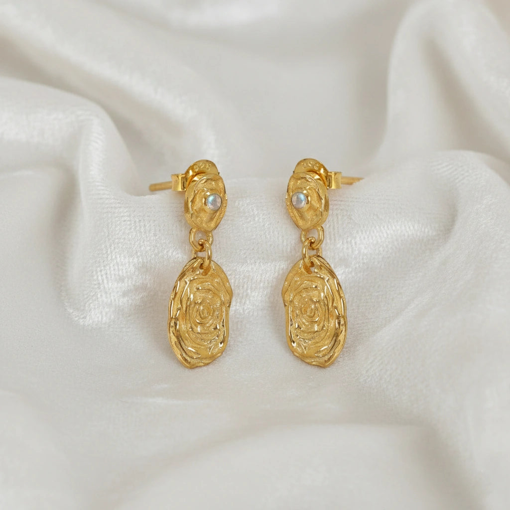 Gold_dangle_earrings_with_gold_plated_silver_textured_oval_charms_and_moonstone_at_side_angle_on_soft_fabric_background