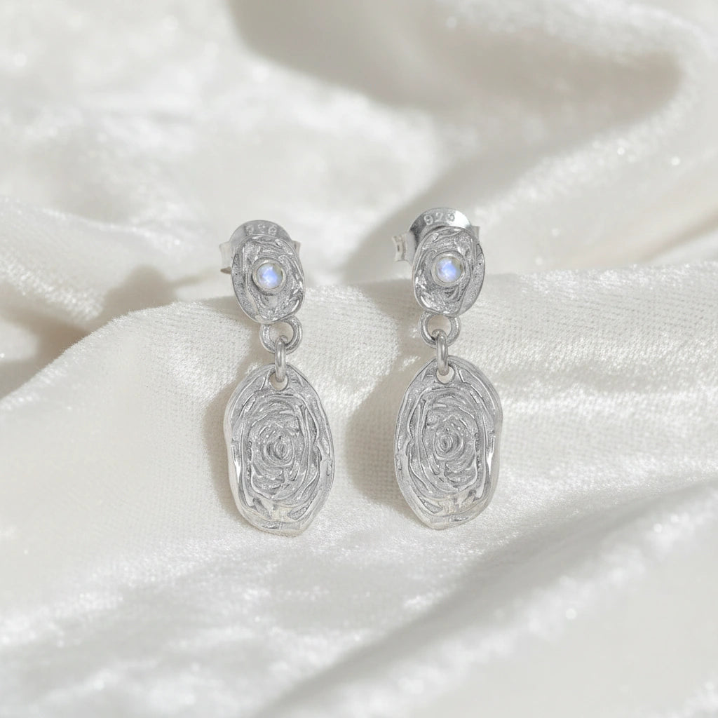 Gold_dangle_earrings_with_sterling_silver_textured_oval_charms_and_moonstone_at_front_angle_on_soft_fabric_background