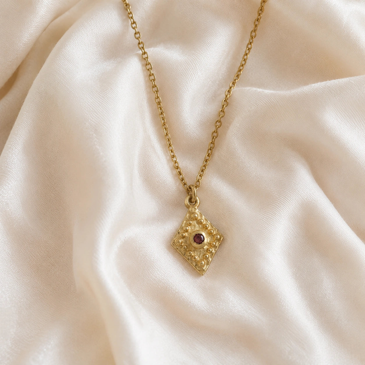 Gold_locket_necklace_gold_plated_silver_front_angle_with_textured_diamond_pendant_and_garnet_center_stone_on_soft_fabric_background