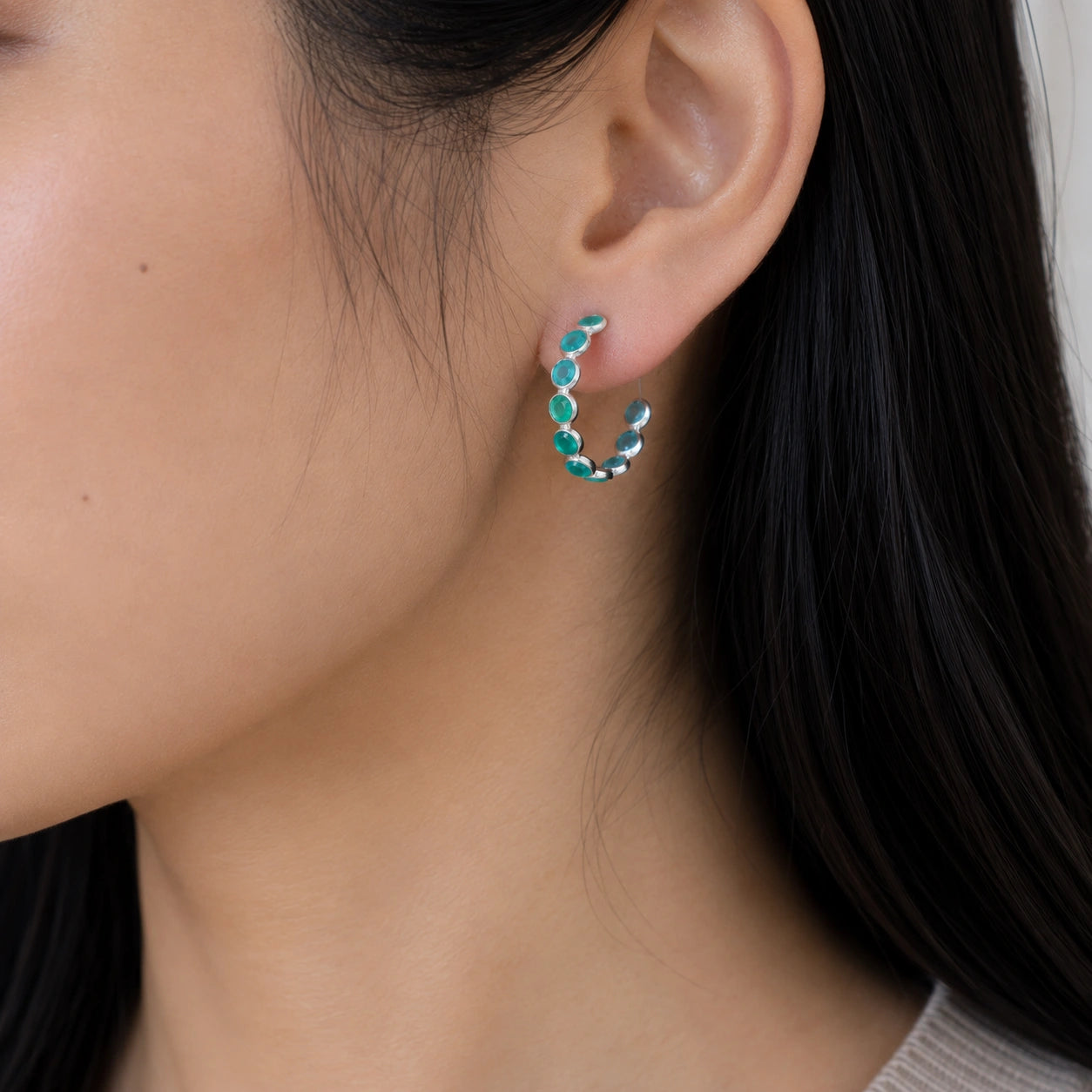 Side angle view of sterling silver hoop earrings featuring green stones worn on the ear background texture