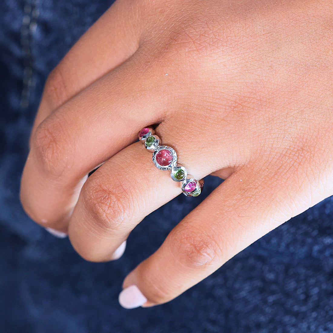 Silver eternity band ring with mixed color gemstones bezel set elevated angle view on soft white velvet fabric background