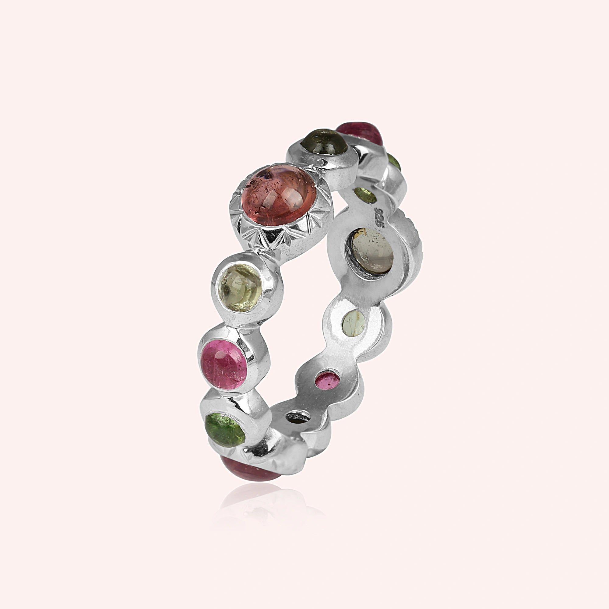 Silver eternity band ring with mixed color gemstones bezel set elevated angle view on solid light pink studio background