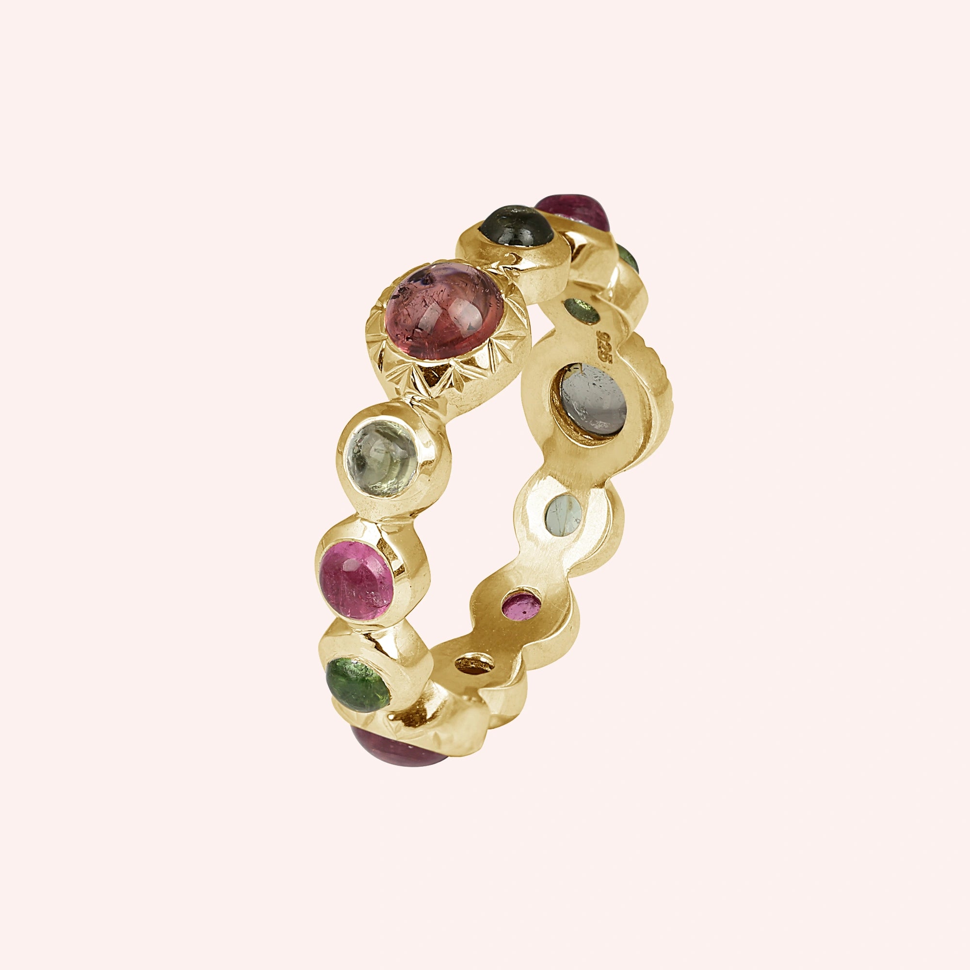 Gold-plated eternity band ring with mixed color tourmaline gemstones elevated view on a solid light pink studio background