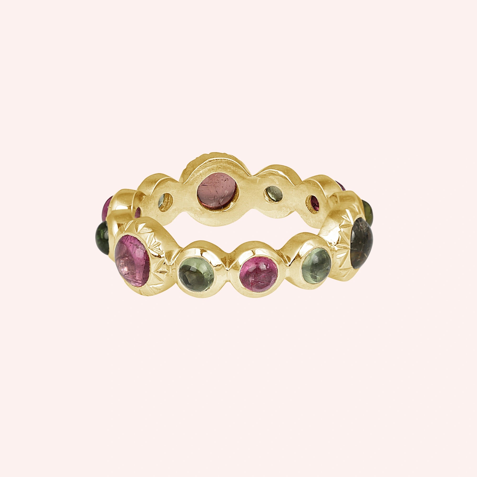 Bezel set gold plated eternity band ring with mixed pink and green tourmaline gemstones straight on view against a light pink background