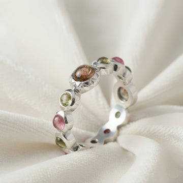 Silver eternity band ring with mixed color gemstones bezel set elevated angle view on soft white velvet fabric background