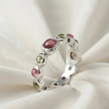 Silver eternity band ring with mixed color gemstones bezel set elevated angle view on soft white velvet fabric background