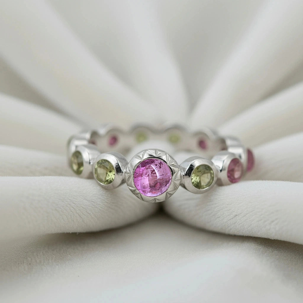 Silver eternity band ring with pink and green tourmaline gemstones bezel set low angle view on a white velvet fabric background
