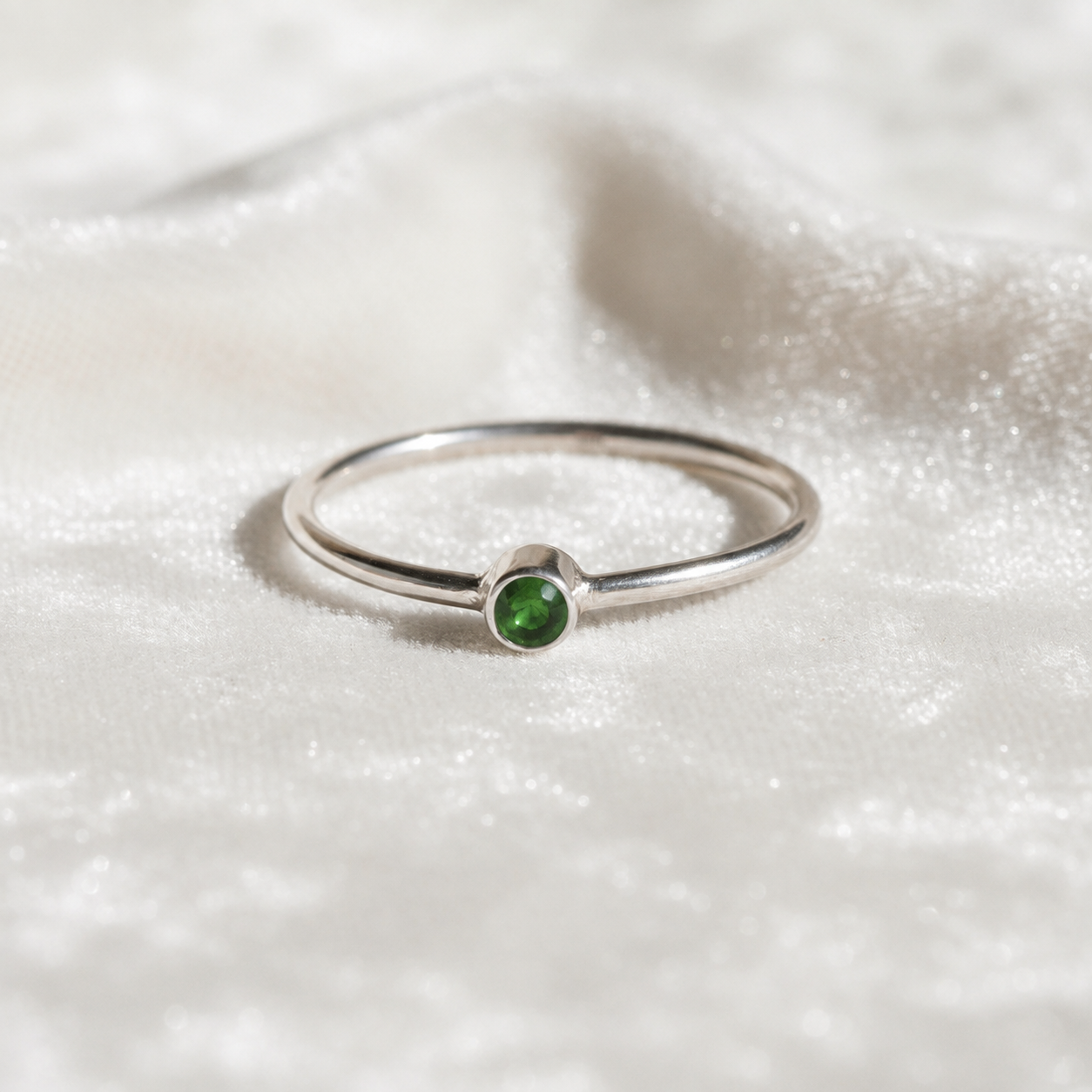 A silver birthstone ring featuring a green tourmaline gemstone, styled as a delicate stacking ring on a soft white fabric background