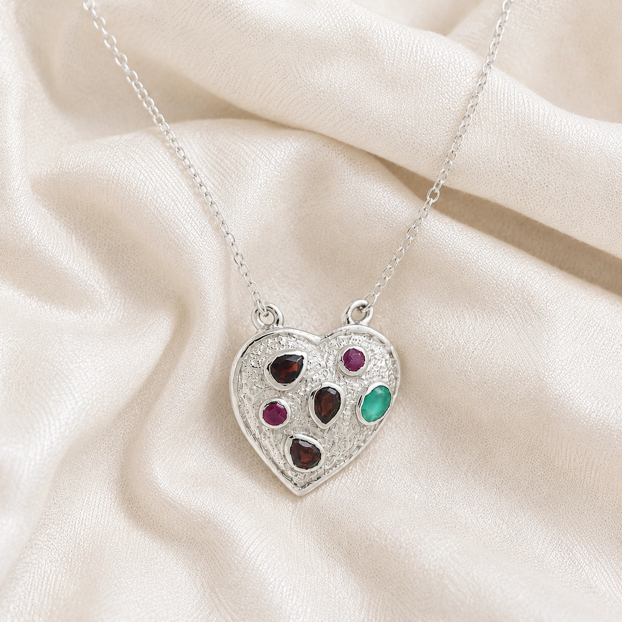 Heart_locket_necklace_sterling_silver_with_garnet_ruby_and_green_onyx_stones_in_slight_angle_view_on_soft_fabric_background