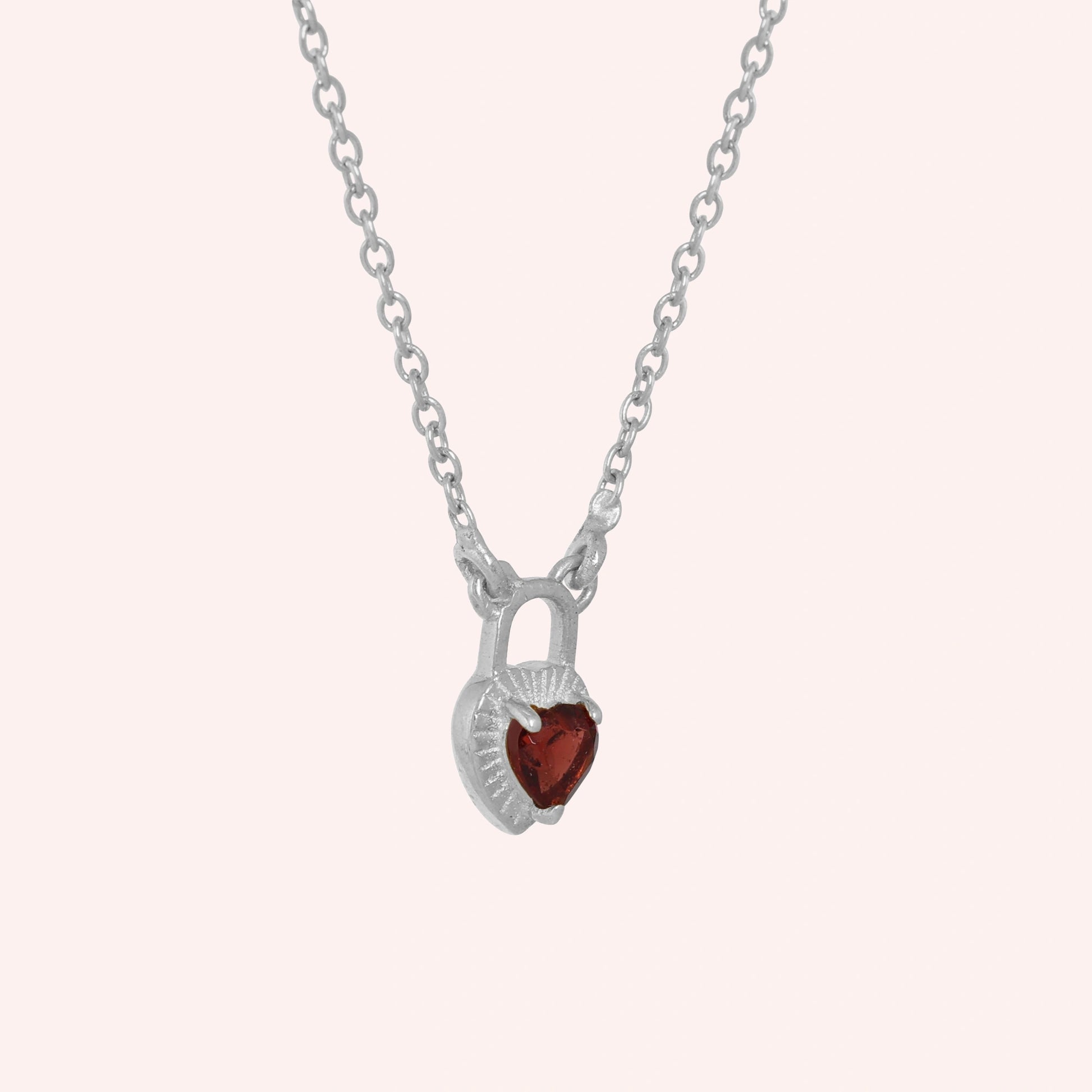 Heart_pendant_necklace_in_sterling_silver_slightly_angled_with_lock_design_and_garnet_stone_charm_on_solid_pink_background