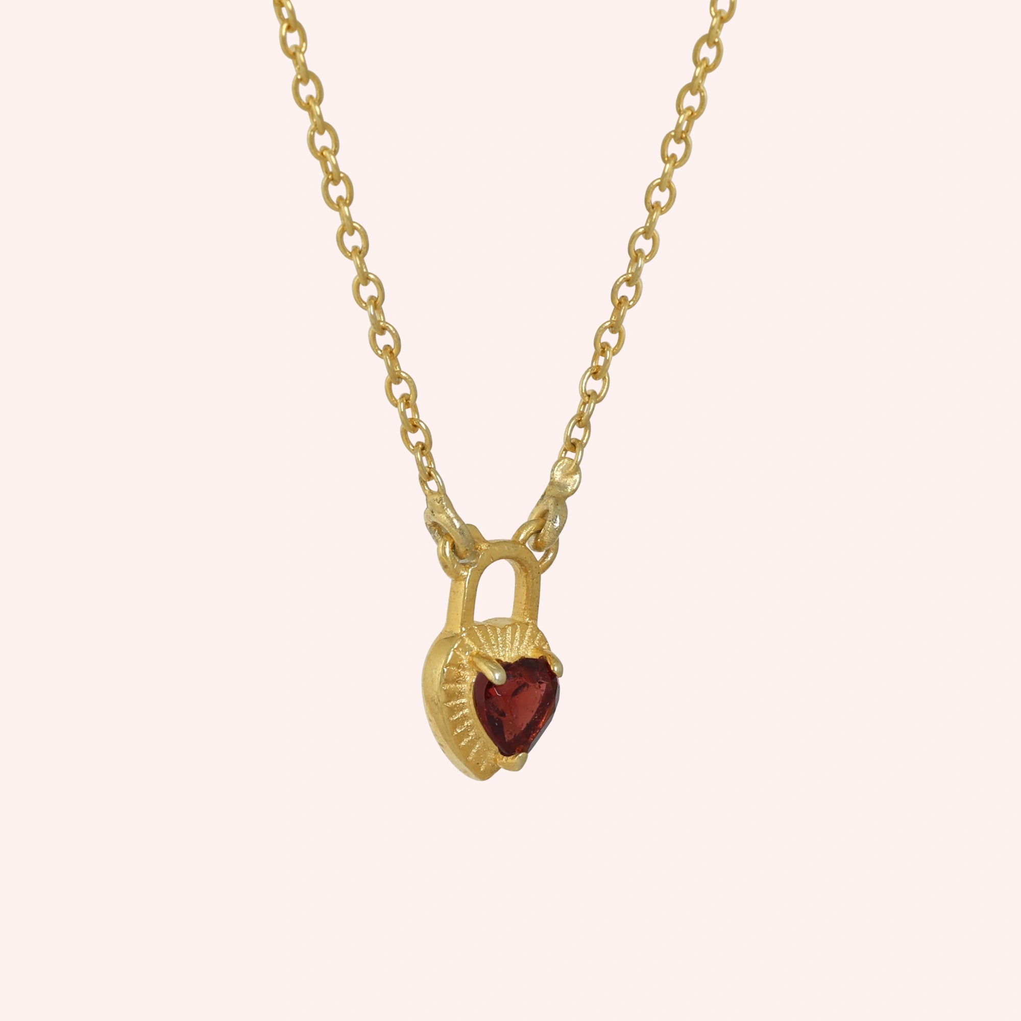 Heart_pendant_necklace_in_gold_plated_silver_slightly_angled_with_lock_design_and_garnet_stone_charm_on_solid_pink_background