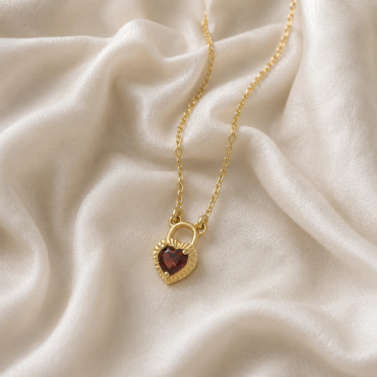 Heart_pendant_necklace_in_gold_plated_silver_slightly_angled_with_lock_design_and_garnet_stone_charm_on_soft_fabric_background