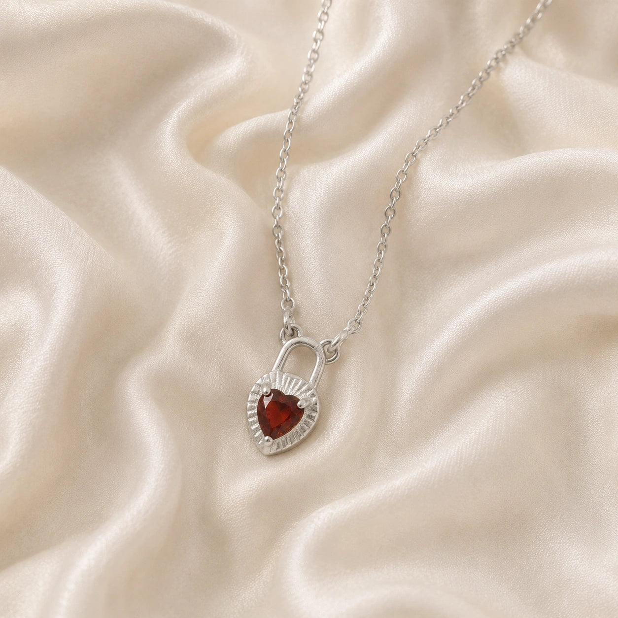 Heart_pendant_necklace_in_sterling_silver_slightly_angled_with_lock_design_and_garnet_stone_charm_on_soft_fabric_background