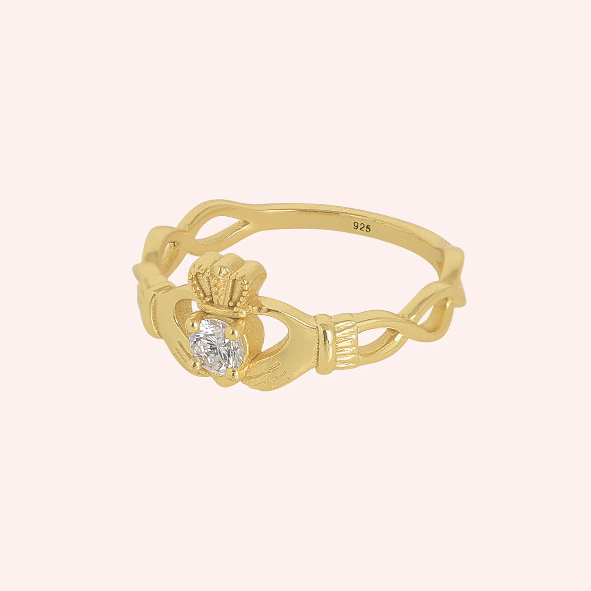 Heart_ring_in_gold_plated_silver_with_a_hand_a_heart_and_a_crown_design_at_side_angle_view_on_solid_pink_background