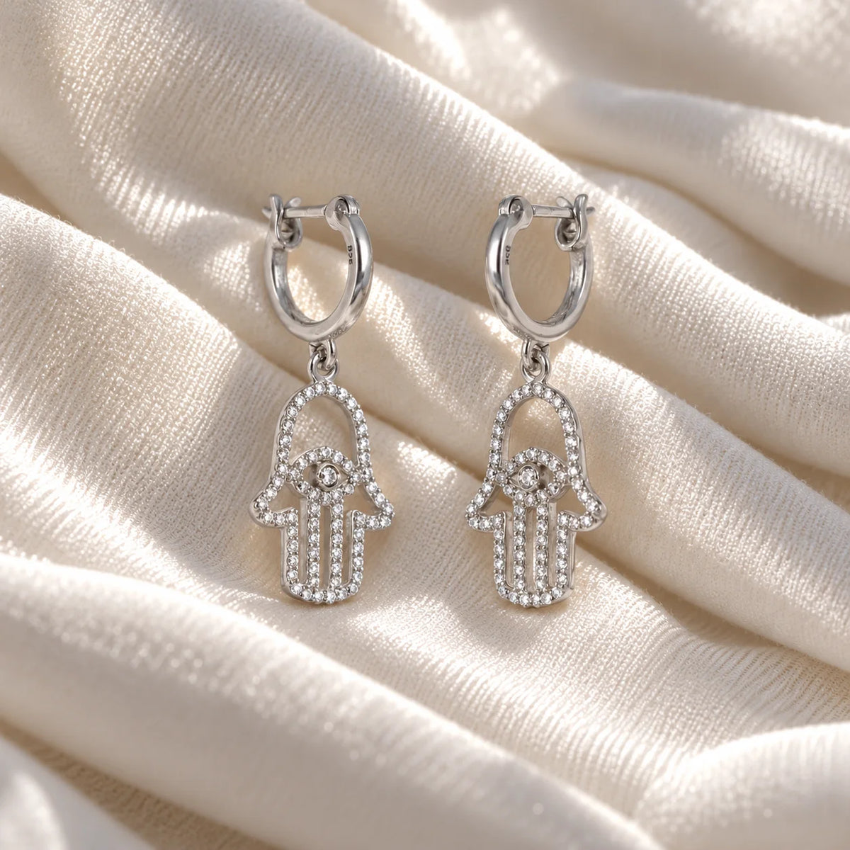 Hoop_earrings_in_sterling_silver_with_a_hamsa_hand_design_at_slight_angle_view_on_soft_fabric_background