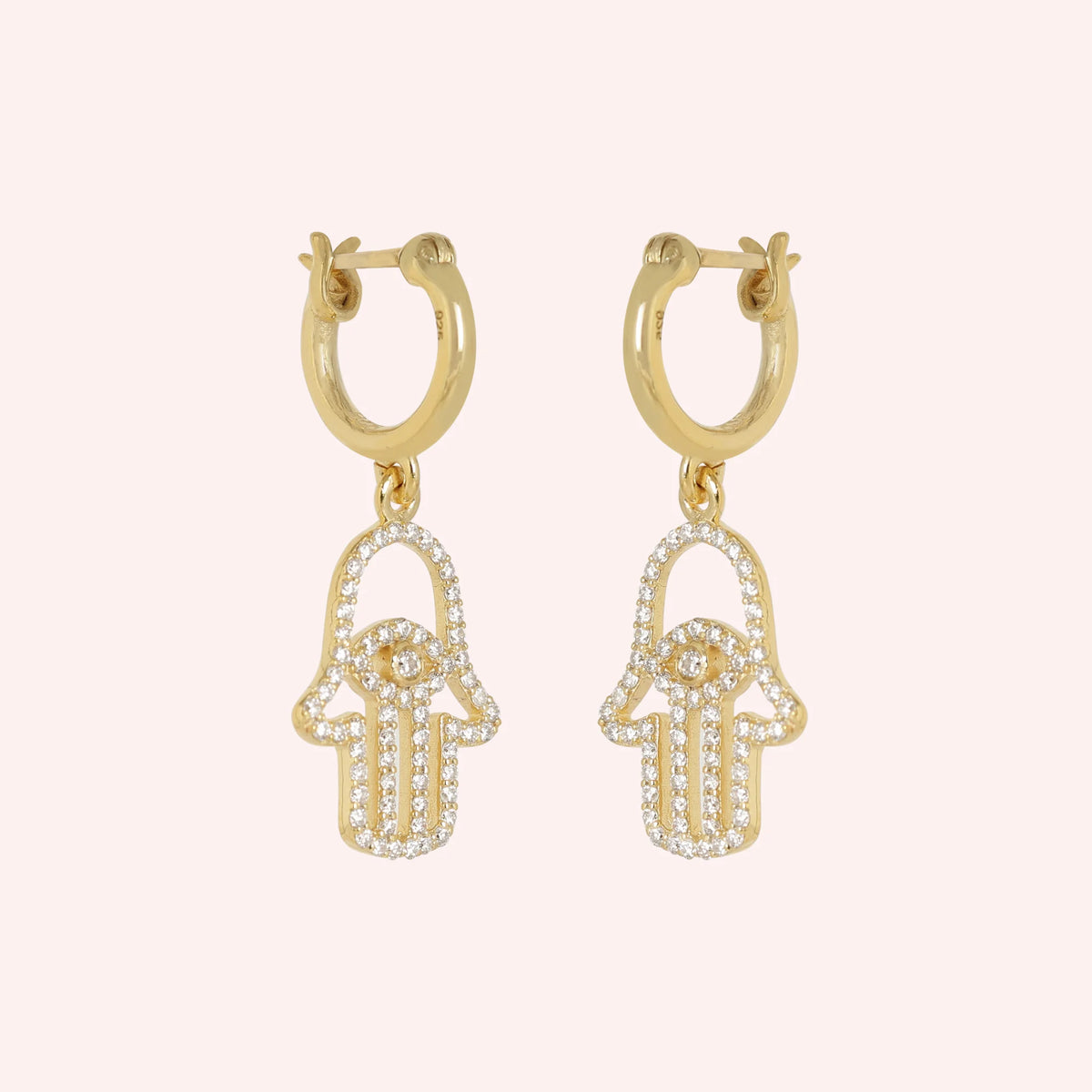 Hoop_earrings_in_gold_plated_silver_with_hamsa_hand_design_and_CZ_stones_at_side_angle_view_on_solid_pink_background