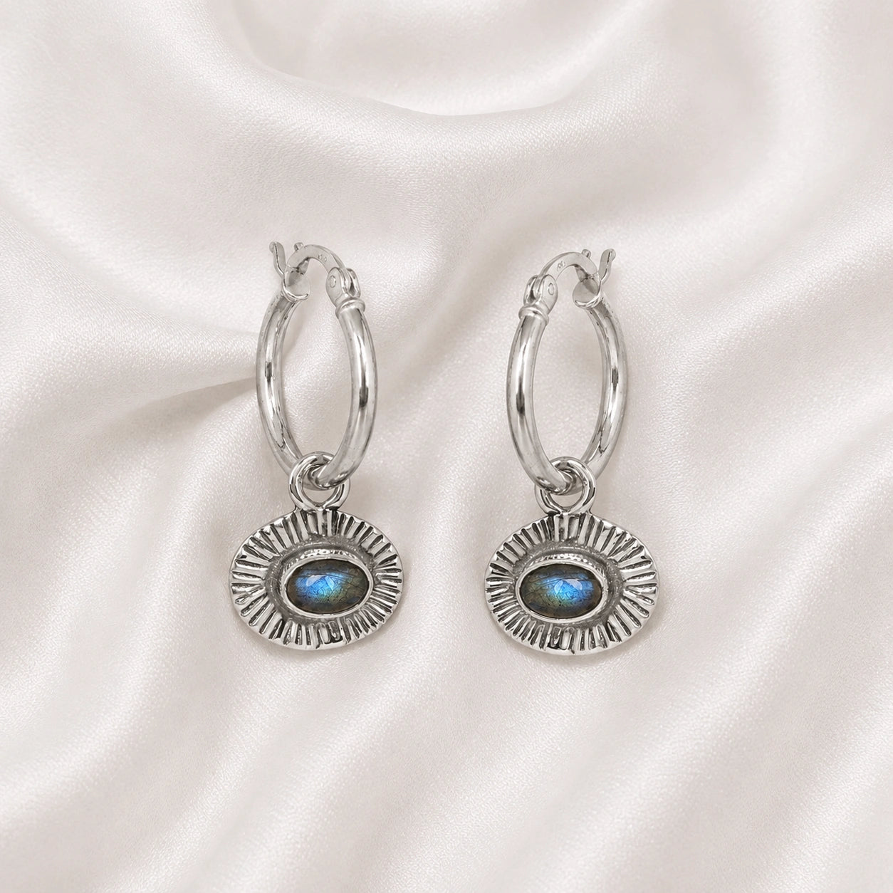 Hoop_earrings_in_sterling_silver_with_oval_labradorite_stone_at_front_angle_view_on_soft_fabric_background