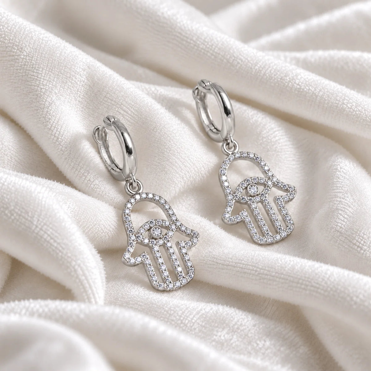 Hoop_earrings_in_sterling_silver_with_hamsa_hand_design_and_CZ_stones_at_side_angle_view_on_soft_fabric_background