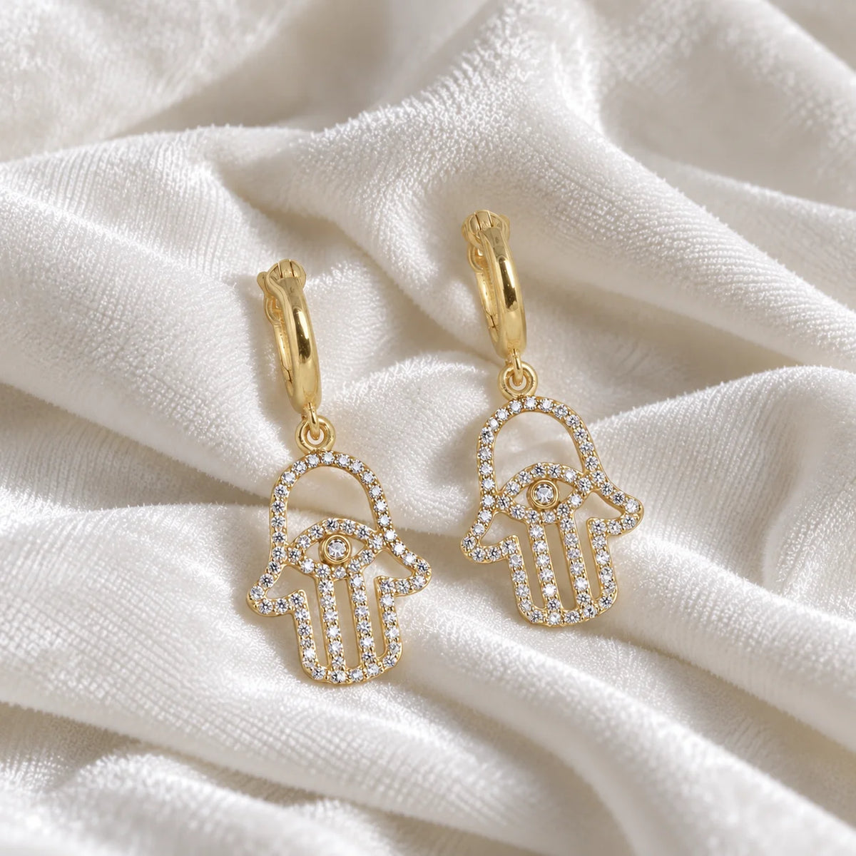 Hoop_earrings_in_gold_plated_silver_with_hamsa_hand_design_and_CZ_stones_at_front_angle_view_on_soft_fabric_background