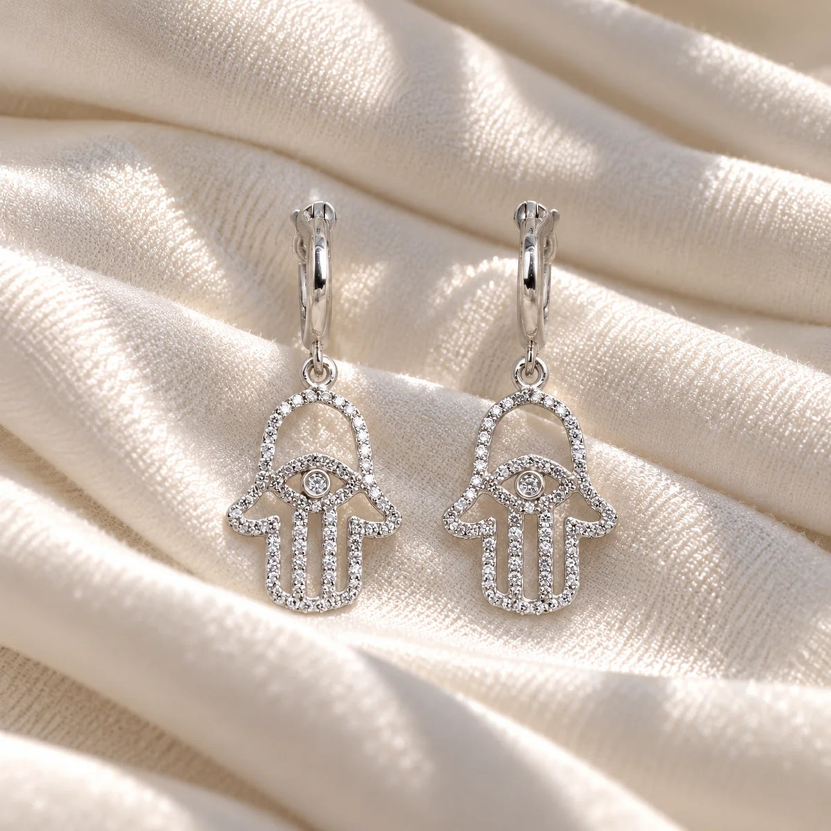 Hoop_earrings_in_sterling_silver_with_a_hamsa_hand_design_at_front_angle_view_on_soft_fabric_background