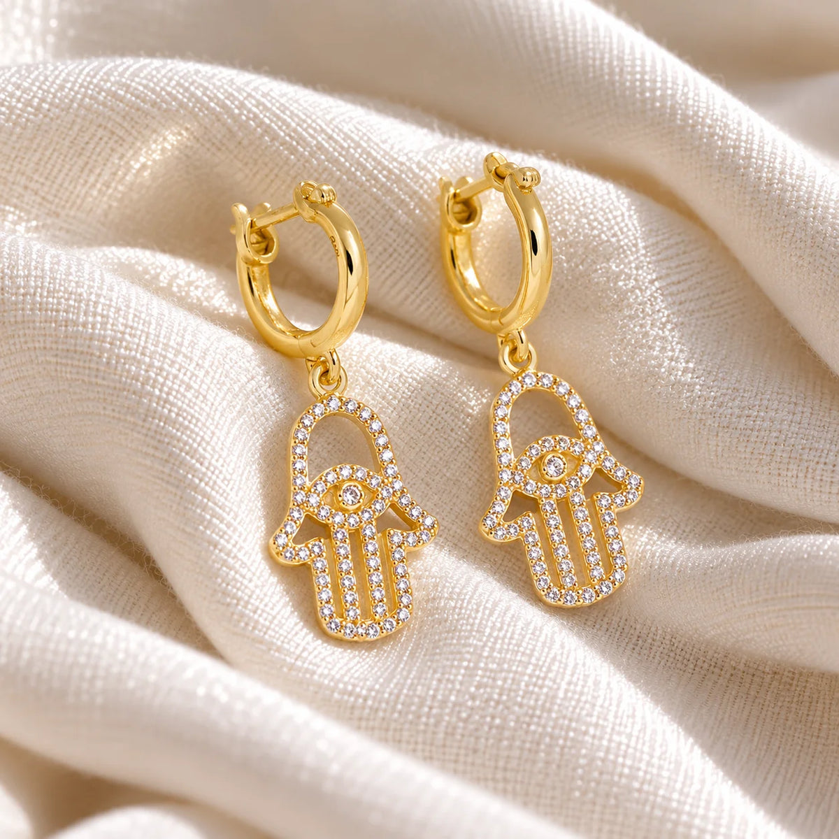 Hoop_earrings_in_gold_plated_silver_with_a_hamsa_hand_design_at_slight_angle_view_on_soft_fabric_background
