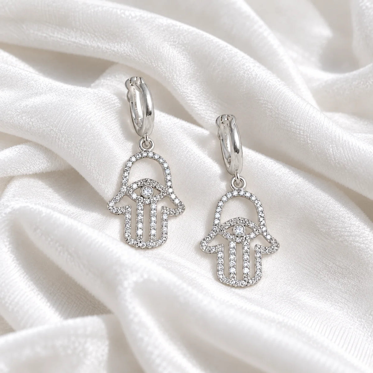 Hoop_earrings_in_sterling_silver_with_hamsa_hand_design_and_CZ_stones_at_front_angle_view_on_soft_fabric_background