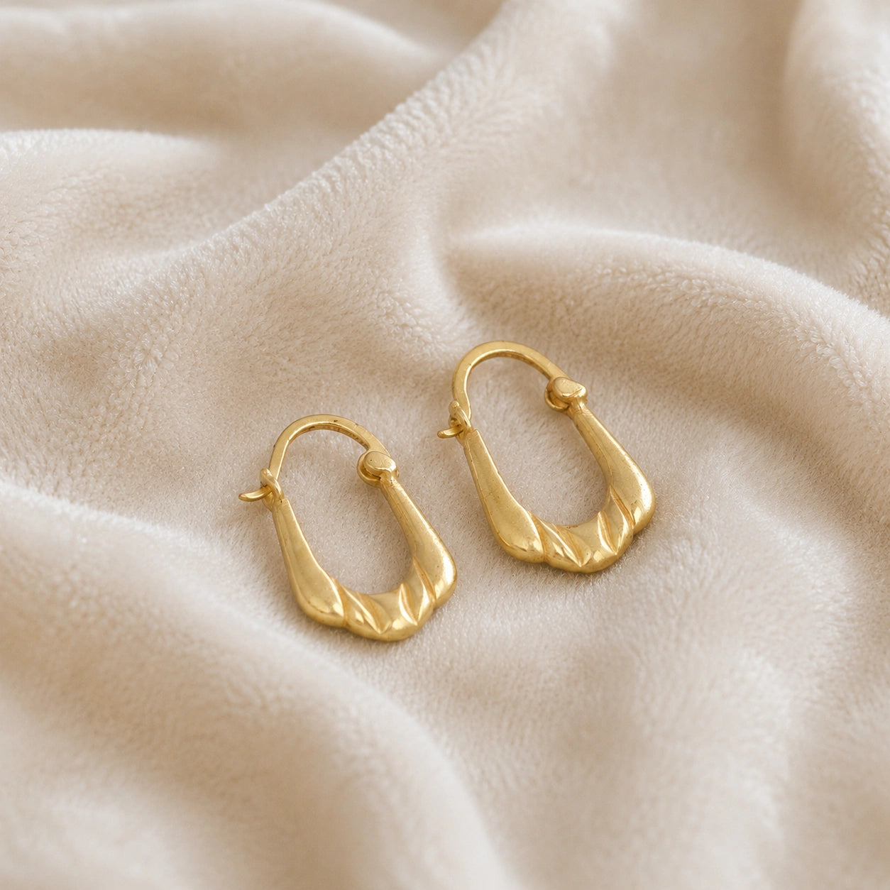Huggie_earrings_in_gold_plated_silver_with_handcrafted_teardrop_hoops_at_front_angle_on_soft_fabric_background