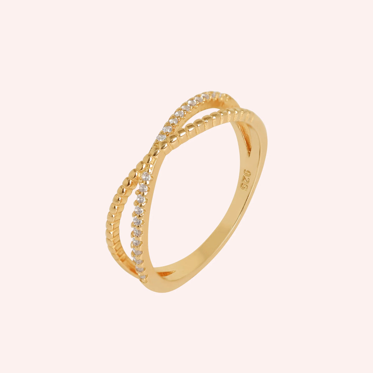 Infinity_ring_in_gold_plated_silver_with_criss_cross_design_and_CZ_stones_at_side_angle_view_on_solid_pink_background