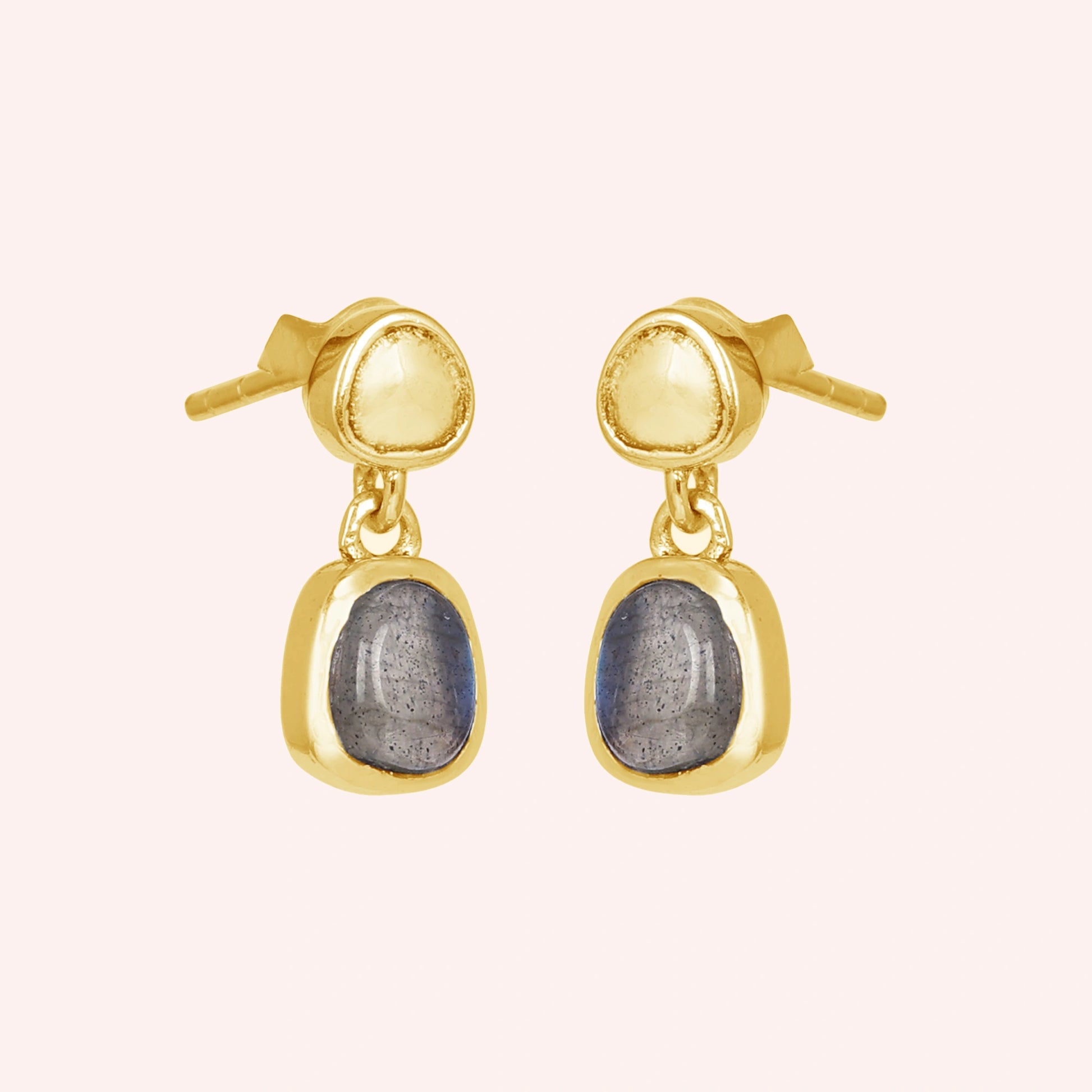 Tiny earrings featuring gold plated studs with labradorite charm stones viewed on a slight angle with light pink surface