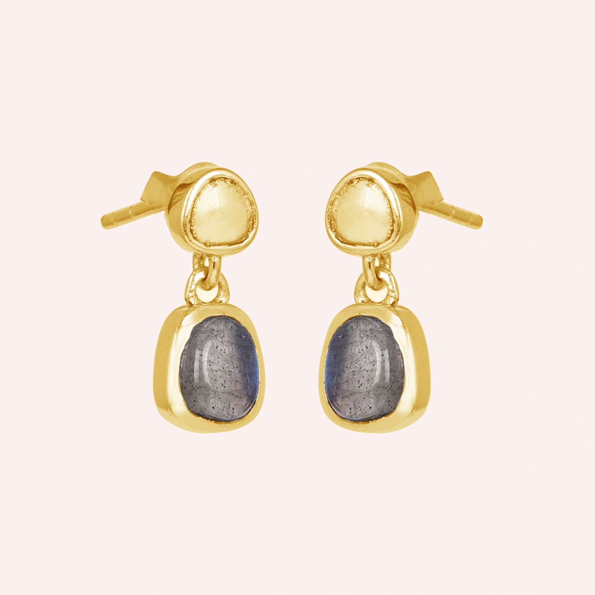 Tiny earrings featuring gold plated studs with labradorite charm stones viewed on a slight angle with light pink surface