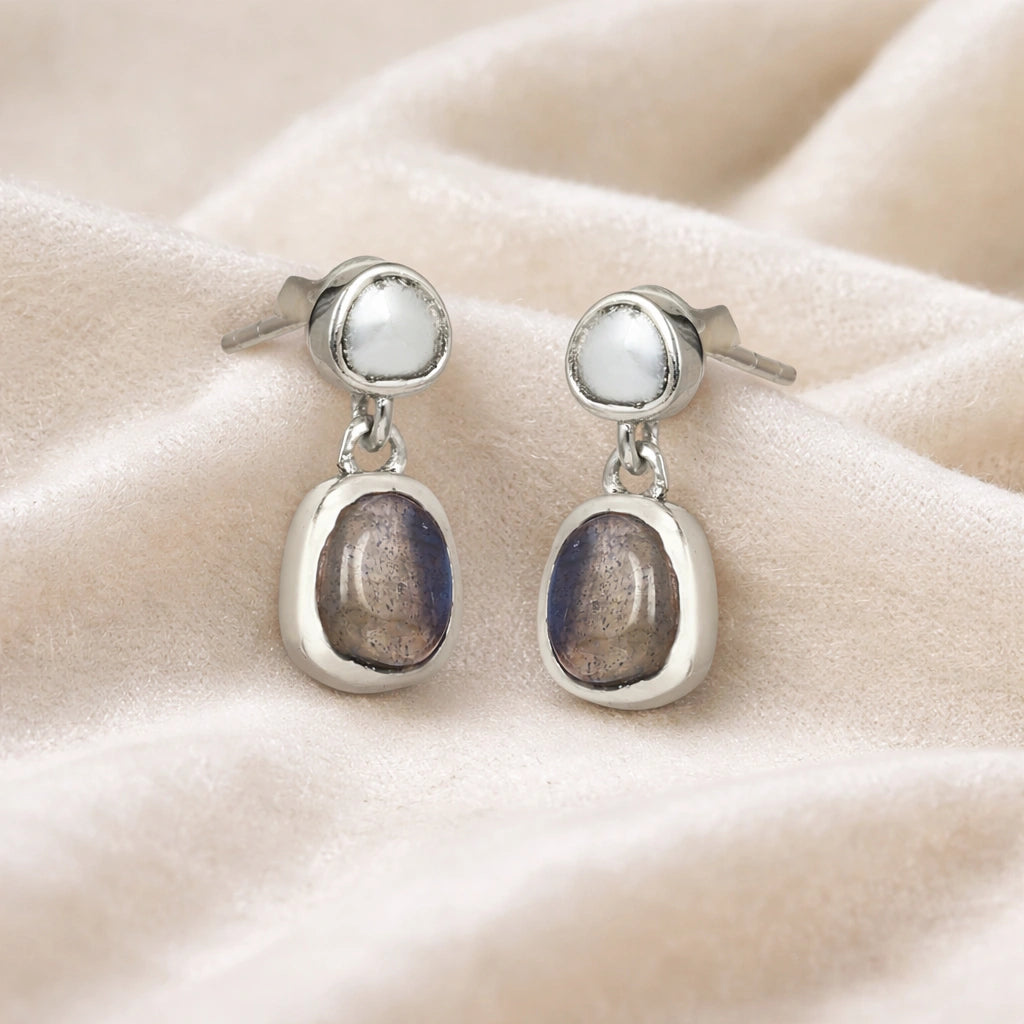 Tiny earrings featuring silver studs with labradorite charm stones viewed straight on a soft white draped fabric